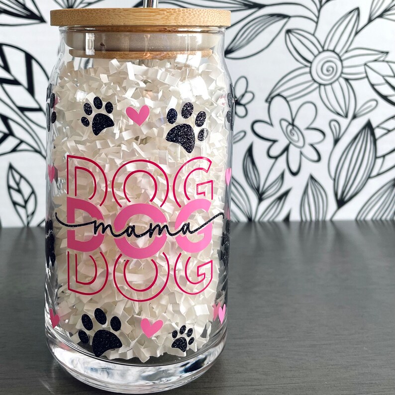 Op de afbeelding: Een heldere glazen beker met een bamboe deksel en rietje, gevuld met wit papier. De beker is versierd met roze tekst met de tekst "DOG mama DOG" en pootafdrukken en hartjes. De achtergrond is een zwart-wit bloemenpatroon.
