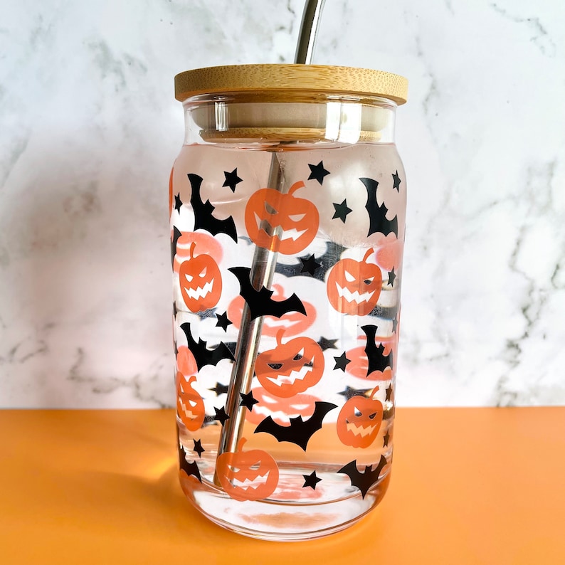 May include: A clear glass reusable water bottle with a bamboo lid and a stainless steel straw. The bottle is decorated with a Halloween themed pattern of orange pumpkins and black bats.