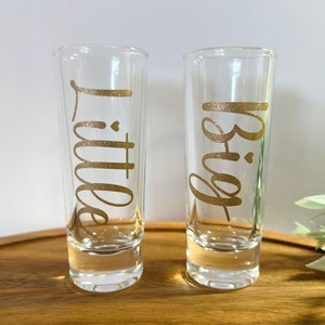 May include: Two clear glass shot glasses with gold glitter lettering. The left glass says "Little" and the right glass says "Big".