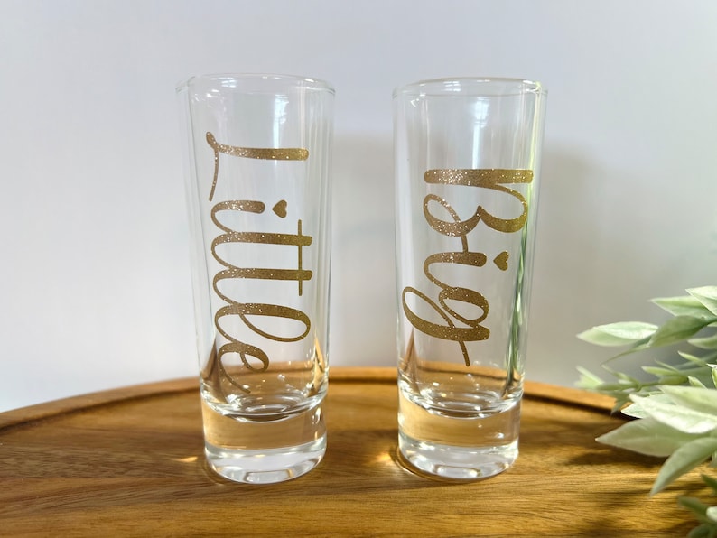 May include: Two clear glass shot glasses with gold glitter lettering. The left glass says "Little" and the right glass says "Big".