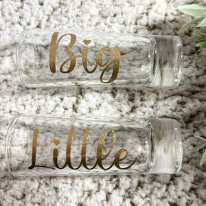 May include: Two clear glass tumblers with gold glitter lettering. The top tumbler says "Big" and the bottom tumbler says "Little".