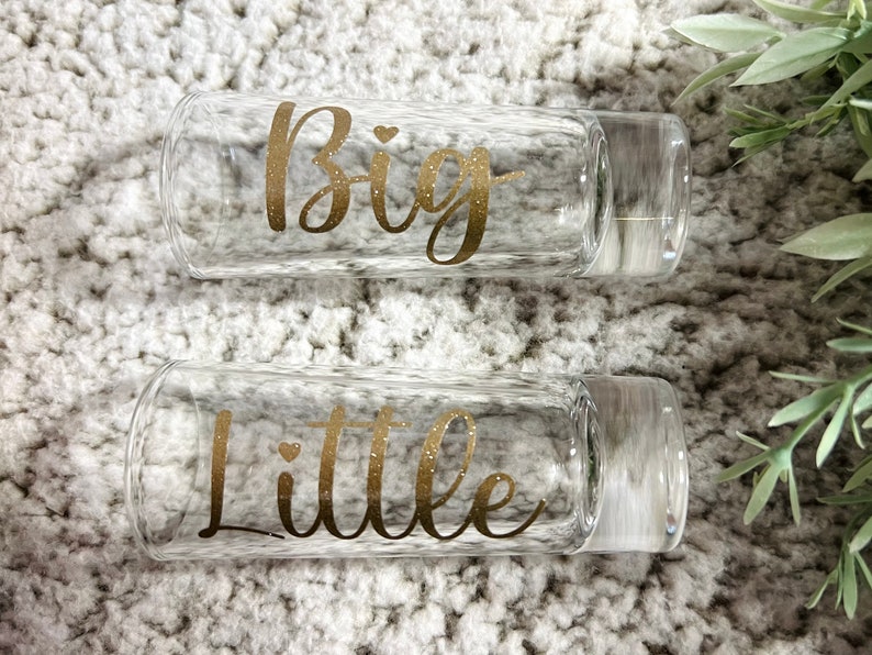 May include: Two clear glass tumblers with gold glitter lettering. The top tumbler says "Big" and the bottom tumbler says "Little".