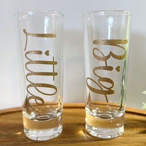 May include: Two clear glass shot glasses with gold glitter lettering. The left glass says "Little" and the right glass says "Big".