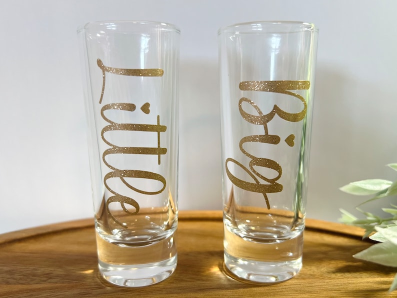 May include: Two clear glass shot glasses with gold glitter lettering. The left glass says "Little" and the right glass says "Big".