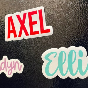May include: Three name stickers on a black surface. The name "Axel" is in red block letters. The name "Ellie" is in teal script, and the name "Jaidyn" is in pink script.