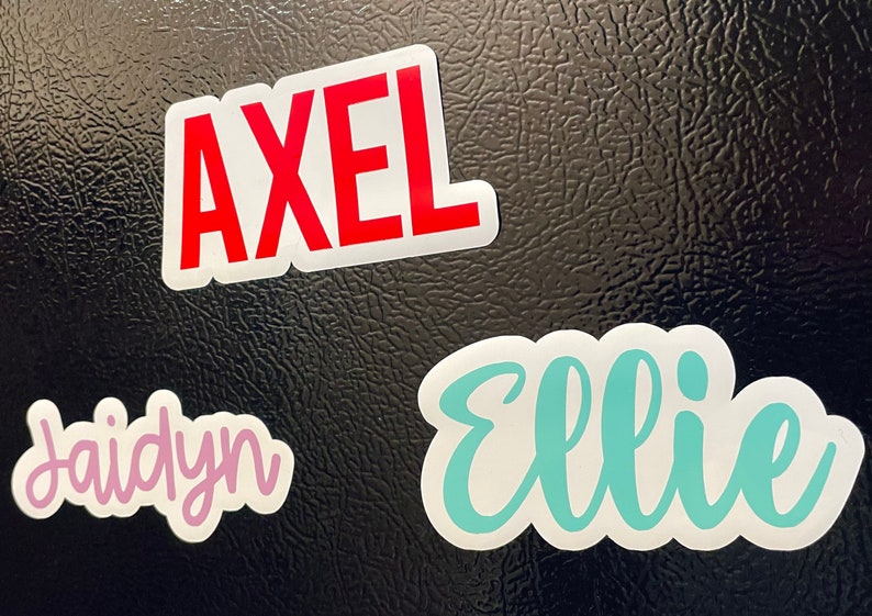 May include: Three name stickers on a black surface. The name "Axel" is in red block letters. The name "Ellie" is in teal script, and the name "Jaidyn" is in pink script.