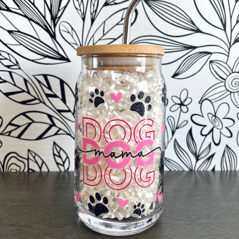 Op de afbeelding: Een heldere glazen beker met een bamboe deksel en een roestvrijstalen rietje. De beker is versierd met roze en zwarte pootafdrukken en de woorden "DOG mama DOG". De beker is gevuld met witte papiersnippers.