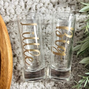 May include: Two clear glass shot glasses with gold lettering. One glass says "Big" and the other says "Little".