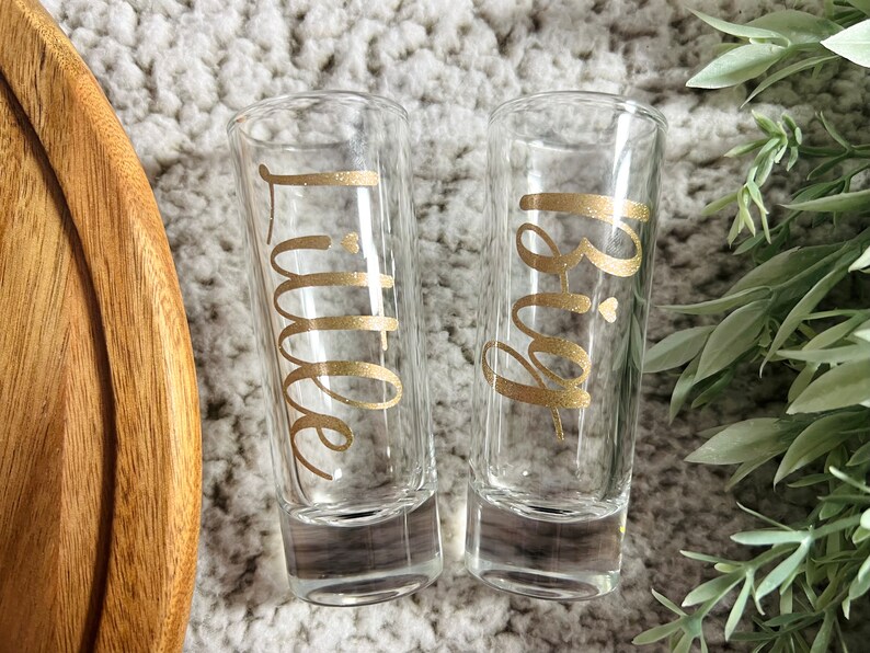 May include: Two clear glass shot glasses with gold lettering. One glass says "Big" and the other says "Little".