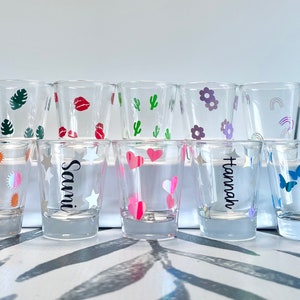 May include: Ten clear glass shot glasses with various colorful designs, including hearts, stars, butterflies, rainbows, flowers, lips, and cacti. Some glasses have names written on them, including "Sami" and "Hannah".