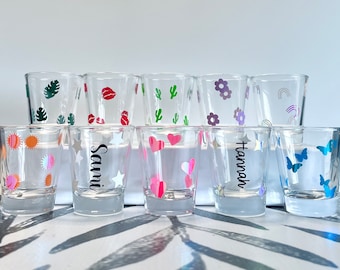 May include: Ten clear glass shot glasses with various colorful designs, including hearts, stars, butterflies, rainbows, flowers, lips, and cacti. Some glasses have names written on them, including "Sami" and "Hannah".
