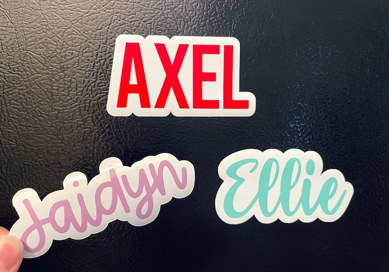 May include: Three name stickers: "Axel" in red block letters, "Jaidyn" in pink script, and "Ellie" in turquoise script. All names are on white backgrounds. The stickers are displayed on a textured, dark surface.