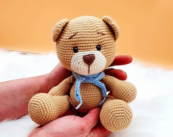 May include: A crocheted brown teddy bear with a blue scarf, sitting in the hands of a person.