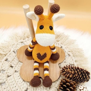 Op de afbeelding: Een gehaakt giraffen speelgoed met bruine en oranje strepen, bruine oren en een bruin hart op de borst. De giraffe zit op een houten standaard met een bloemvormige basis.