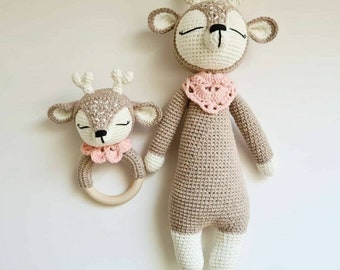 May include: Two crocheted deer toys. The larger deer is standing and has a pink crocheted bib. The smaller deer is attached to a wooden ring and has a pink crocheted collar.