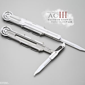 Peut inclure: Deux paires de lames cachées Haytham Kenway argentées. Les lames ont un mécanisme en forme de roue dentée et une lame pointue. Le texte "AC III HAYTHAM KENWAY HIDDEN BLADE PAIR" est visible.