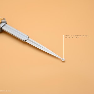 May include: A silver and gold folding knife with a small removable safety tab. The knife is closed and lying on a light brown surface.
