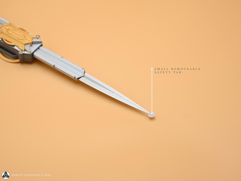 May include: A silver and gold folding knife with a small removable safety tab. The knife is closed and lying on a light brown surface.