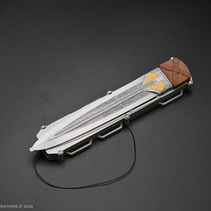 K&ouml;nnte beinhalten: Ein silbernes Klappmesser mit braunem Griff und goldenen Akzenten. Das Messer hat ein dekoratives Muster auf der Klinge und eine Schnur, die am Griff befestigt ist.