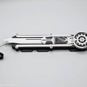 K&ouml;nnte beinhalten: Ein schwarzes und silbernes Klappmesser mit einem zahnradf&ouml;rmigen Design am Griff. Das Messer hat eine silberne Klinge und eine schwarze Schnur, die am Griff befestigt ist.