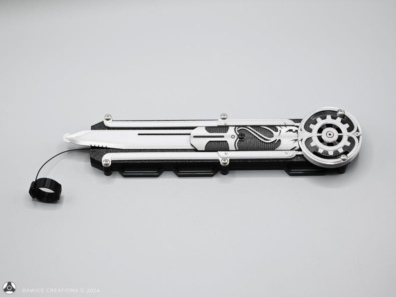 K&ouml;nnte beinhalten: Ein schwarzes und silbernes Klappmesser mit einem zahnradf&ouml;rmigen Design am Griff. Das Messer hat eine silberne Klinge und eine schwarze Schnur, die am Griff befestigt ist.