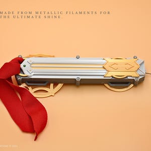 May include: A silver and gold-colored metal object with a red ribbon attached. The object has a textured surface and is made from metallic filaments. The text "MADE FROM METALLIC FILAMENTS FOR THE ULTIMATE SHINE." is visible above the object.