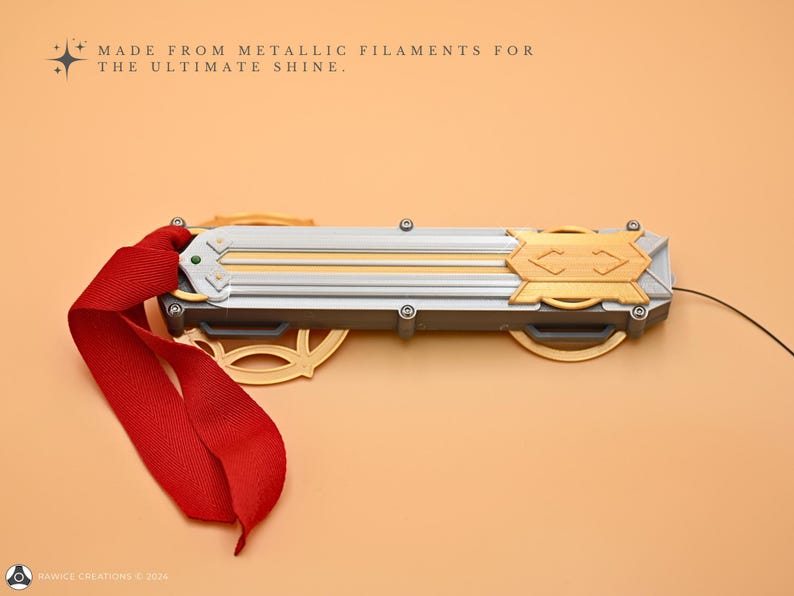 May include: A silver and gold-colored metal object with a red ribbon attached. The object has a textured surface and is made from metallic filaments. The text "MADE FROM METALLIC FILAMENTS FOR THE ULTIMATE SHINE." is visible above the object.