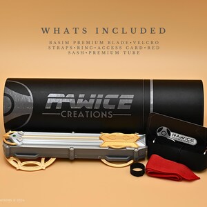 May include: A black tube with the text "Rawice Creations" printed on it. The tube contains a silver and gold blade with a green gem. The image also shows a black strap, a red sash, and a card with the text "Rawice Creations".