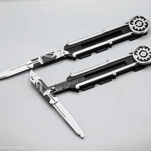 K&ouml;nnte beinhalten: Zwei schwarze und silberne Klappmesser mit einem Zahnrad-Design am Griff. Die Messer haben eine silberne Klinge mit einem schwarz-wei&szlig;en Muster.