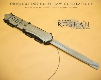 May include: A silver-colored, retractable blade weapon with a black handle and a black and silver design. The blade is extended and the weapon is lying on a light orange surface. The text "AC MIRAGE ROSHAN HIDDEN BLADE" is printed on the surface.