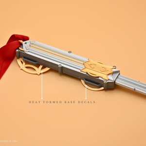 May include: A silver and gold prop sword with a red ribbon attached to the handle. The sword has a detailed design with a gold emblem and the words "HEAT FORMED BASE DECALS".
