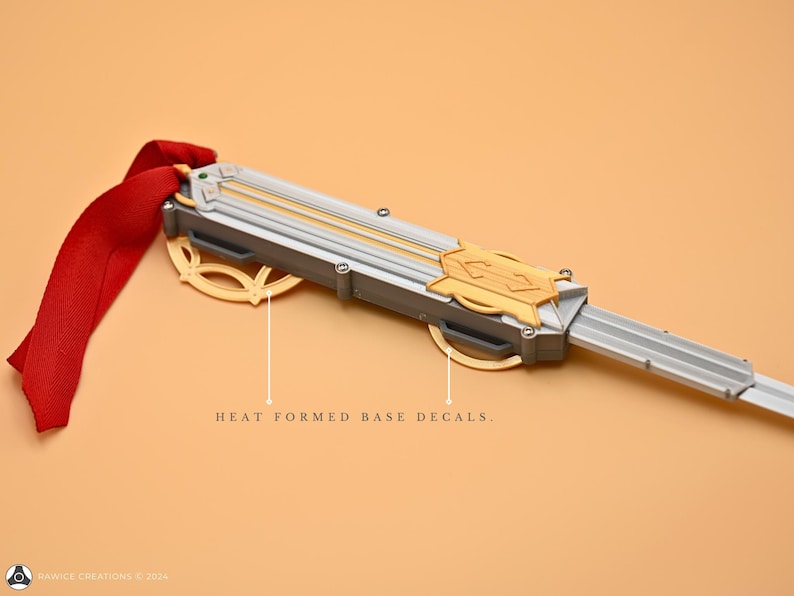 May include: A silver and gold prop sword with a red ribbon attached to the handle. The sword has a detailed design with a gold emblem and the words "HEAT FORMED BASE DECALS".