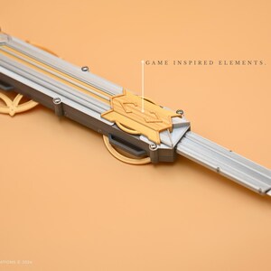 May include: A silver and gold-colored prop weapon with a blade and a decorative handle. The handle features a gold design with a geometric pattern. The text "GAME INSPIRED ELEMENTS." is visible in the image.