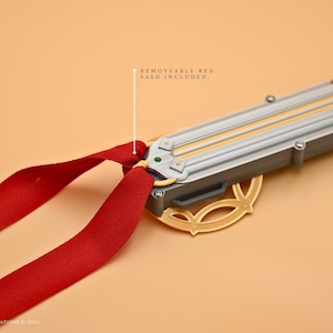 May include: A gold and silver decorative sword with a red sash. The sword has a detailed design and is attached to a red ribbon.