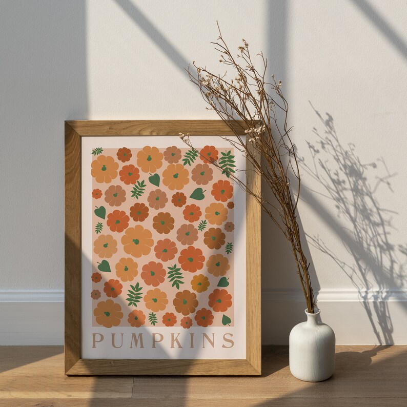 May include: A framed print featuring a pattern of orange and brown pumpkins on a pale pink background. The print has the word "PUMPKINS" in brown text at the bottom. The frame is brown wood and the print is hanging on a white wall.