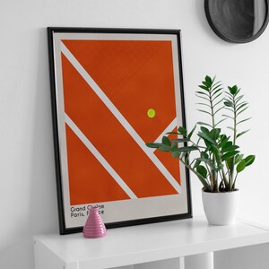 May include: A framed poster with a minimalist design of a tennis court in orange and white with a yellow tennis ball. The poster has the text "Grand Chelem Paris, France" at the bottom.