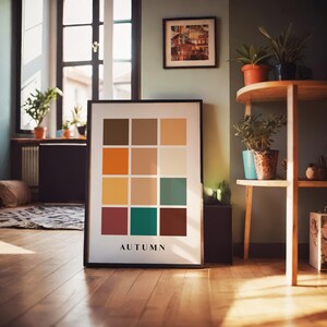May include: A framed print with a grid of 12 colorful squares, labeled "AUTUMN". The squares are in shades of brown, orange, yellow, green, and red. The print is on a wooden floor in a room with plants and a window.