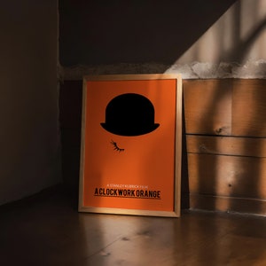 Puede incluir: Un p&oacute;ster enmarcado con fondo naranja y una silueta negra de un bomb&iacute;n con un ojo. El texto "A Stanley Kubrick Film A Clockwork Orange" est&aacute; impreso en negro en la parte inferior del p&oacute;ster.