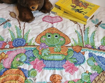 Pode incluir: Um cobertor acolchoado branco com um desenho de ponto cruz de um sapo e tartarugas. O cobertor é decorado com um coração rosa e um urso de peluche castanho.
