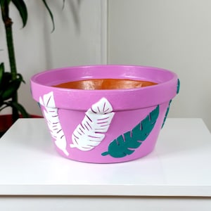 Könnte beinhalten: Ein rosafarbener Terrakotta-Pflanztopf mit weißen und grünen bemalten Blättern. Der Topf ist mit einem tropischen Blattdesign verziert.