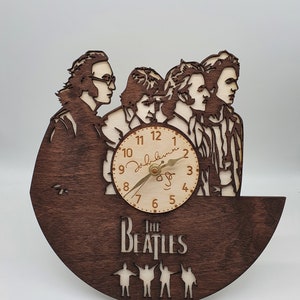 May include: A wooden clock shaped like a record with a clock face featuring the Beatles band members. The clock face has Roman numerals and a signature that reads "John Lennon". The clock is brown and has a stand.