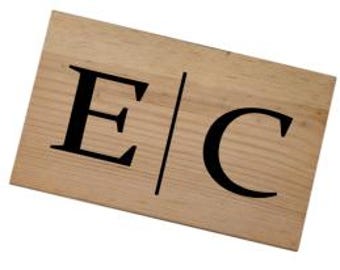 Può includere: Cartello in legno con le lettere "E" e "C" in nero, separate da una linea verticale. Il cartello presenta una venatura naturale del legno.
