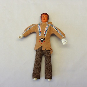 May include: A vintage doll dressed in a Native American outfit. The doll has a tan suede-like shirt with fringe and decorative blue and red patterns. The doll's pants are brown with a textured pattern. The doll has white hands and feet.