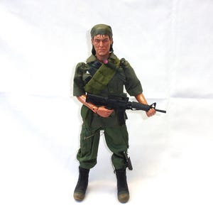 May include: A military action figure in a green uniform, holding a black rifle. The figure wears a green headscarf and black boots. A green strap is across the chest. The figure is standing on a white surface.