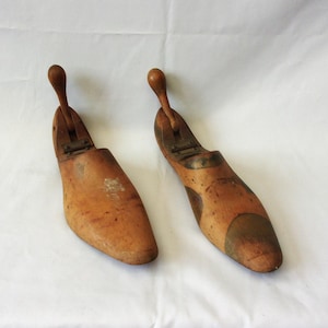 May include: Two antique wooden shoe forms with a light brown finish. Each form has a rounded toe and a wooden handle. The shoe forms are displayed on a white background.