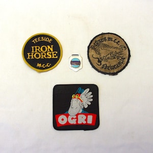 May include: A collection of vintage patches and pins. Includes a round patch with "TEESIDE IRON HORSE M.C.C." text, a small pin, a round patch with a dragon, and a square patch with "OGRI" text and a cartoon character.