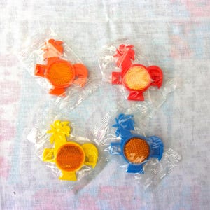 Op de afbeelding: Vier plastic haanreflectoren in oranje, rood, geel en blauw, elk met een oranje reflecterend midden. De reflectoren zijn verpakt in helder plastic. Vintage ontwerp.