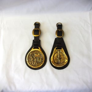 Puede incluir: Dos llaveros de cuero con medallones de latón. Un medallón presenta un grupo de personas caminando, el otro presenta una espada. Ambos medallones tienen una correa de cuero negro.