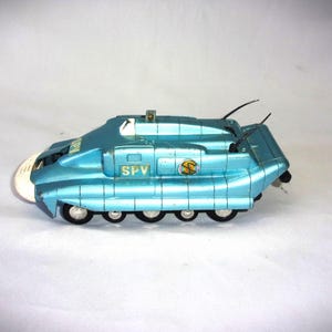 Op de afbeelding: Een vintage, turquoise speelgoedvoertuig met de tekst "SPV" op de zijkant. Het speelgoed heeft een afgeronde voorkant, zes wielen en twee zwarte antennes. Een geel en oranje embleem is ook zichtbaar.