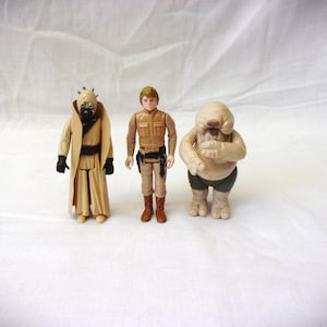 Può includere: Tre action figure di Star Wars: una figura con una tunica marrone e una testa cornuta, una figura color beige con una cintura marrone e un'uniforme marrone e beige, e una figura color beige con una testa rotonda e una fascia verde.